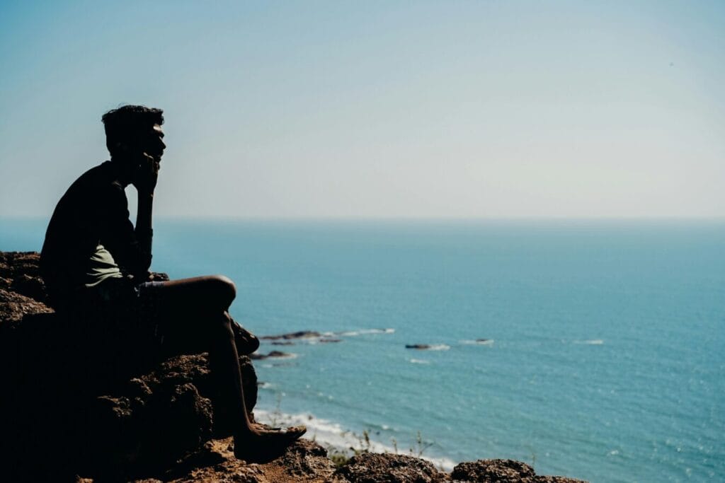how to grow in faith - silhouette of a man sitting on a cliff overlooking a calm ocean on a sunny day.