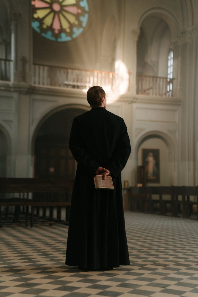 A priest in traditional attire stands in a cathedral interior, holding a book, under colorful stained glass light.